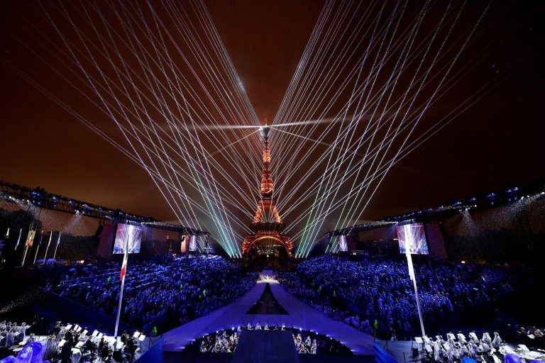 View of the Paris Olympics 2024 Opening Ceremony, showcasing a vibrant blend of lights, art, and culture with iconic Parisian landmarks illuminated against the night sky. Athletes and performers take center stage, celebrating global unity and the rich cultural heritage of France.