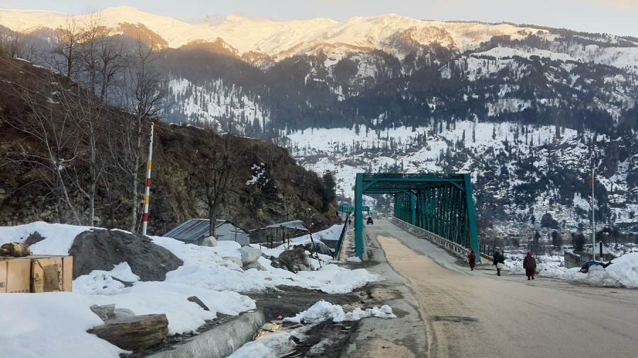 A breathtaking winter scene of Manali, Himachal Pradesh, showcasing snow-covered mountains, frost-kissed pine forests, charming wooden cottages with smoking chimneys, and a glistening river winding through the valley under a crisp blue sky.