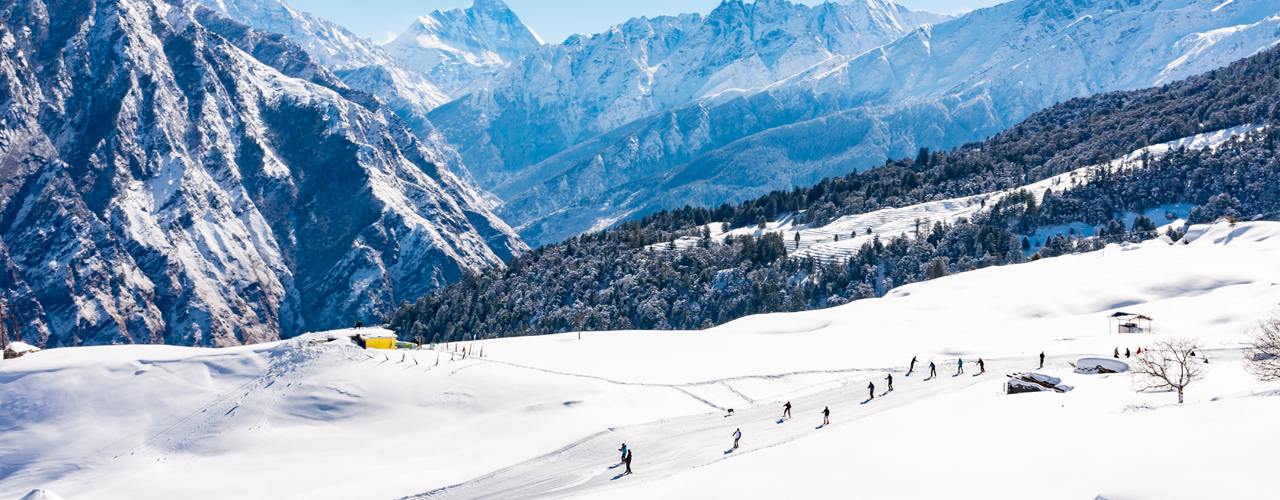 A spectacular winter panorama of Auli, Uttarakhand, featuring pristine snow-covered slopes surrounded by towering Himalayan peaks. Skiers glide down the well-groomed trails, while cable cars soar above offering breathtaking views. Quaint wooden lodges and alpine forests complete the picture of this premier skiing destination.